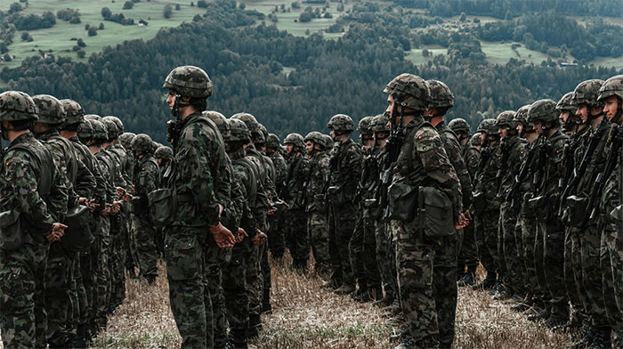 Young soldiers in camouflage standing in formation outdoors with a forested landscape in the background.