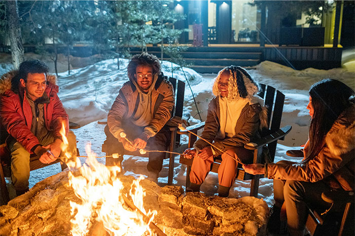Group of young people sitting around a fire pit roasting marshmallows, enjoying a winter evening outdoors together.