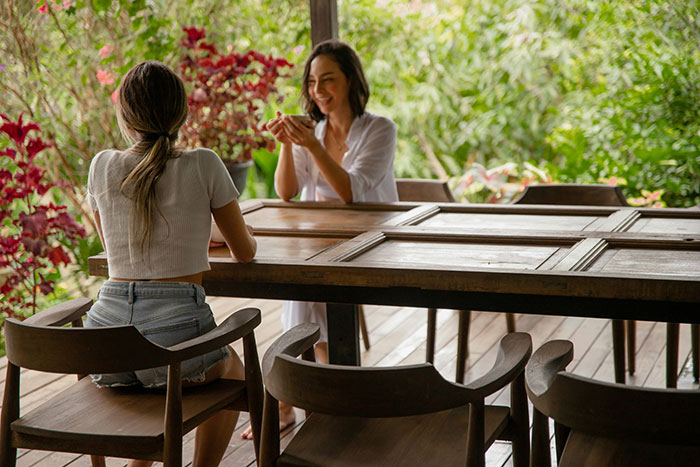 Two women sitting at a wooden table outdoors, one smiling while holding a cup, depicting a fianc&eacute;e and her friend.