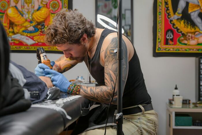 Tattoo artist with sleeve tattoos focused on tattooing a client's arm in a colorful studio setting with Indian art.