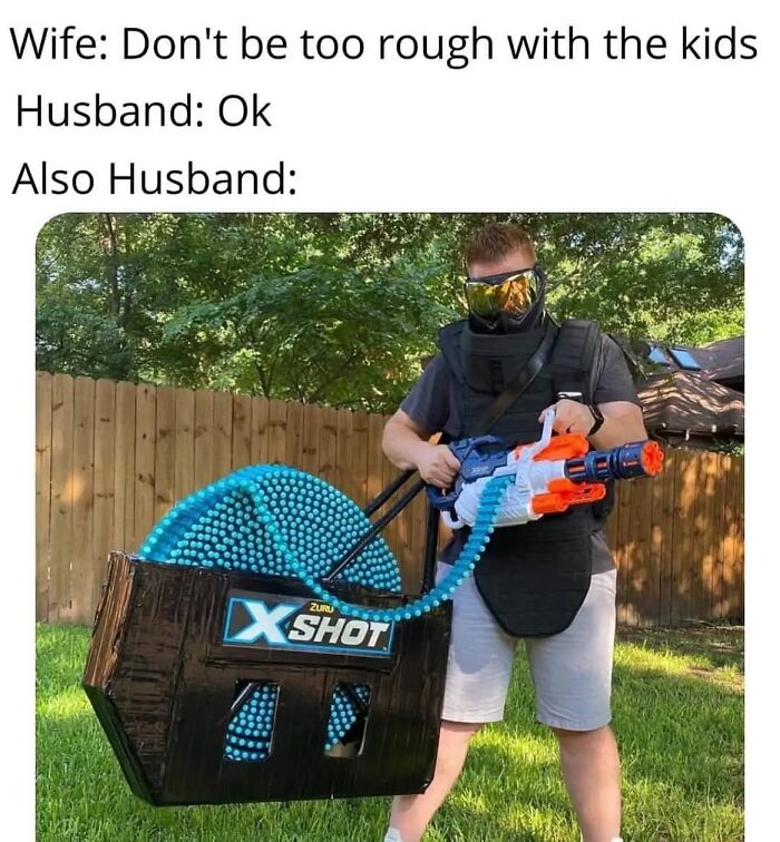 Husband wearing protective gear and holding a large toy gun, humorously depicting chaotic parenting moments.