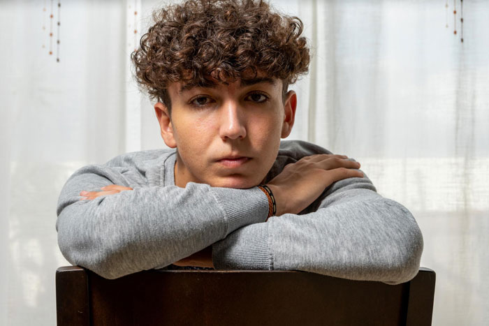 Teenage boy with curly hair sitting with arms crossed on chair, reflecting on toxic parents favoring golden child son.