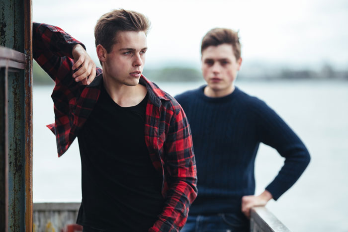 Two young men outdoors by water, one looking upset and distant, depicting tension between favored golden child and younger son.