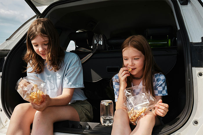 Two young girls sitting in a car trunk eating snacks, illustrating parents sharing baby names they regret giving to kids.