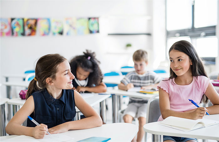 Two girls smiling and writing in notebooks in a bright classroom, illustrating baby names parents regret giving kids.
