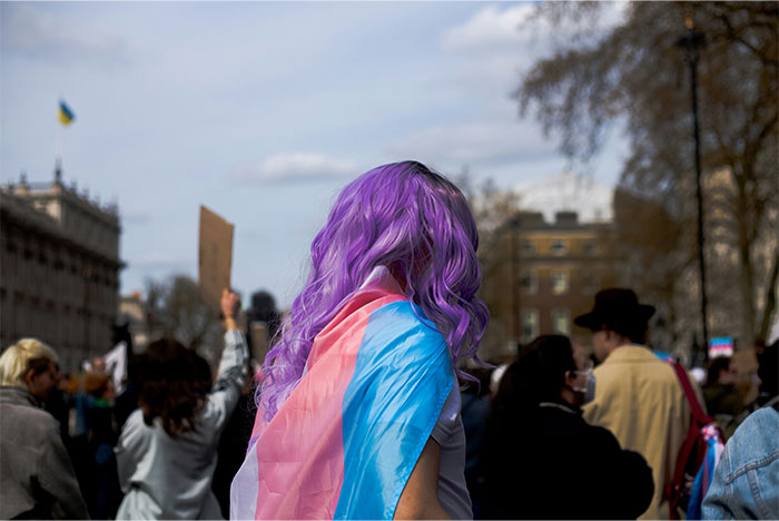 Person with purple hair draped in a transgender pride flag attending a crowd gathering, symbolizing baby names parents regret.