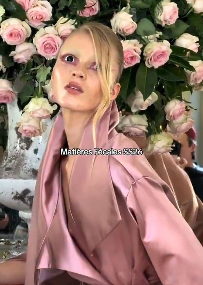 Model with avant-garde makeup posing in pink satin outfit against a backdrop of pink roses at Paris Fashion Week show.