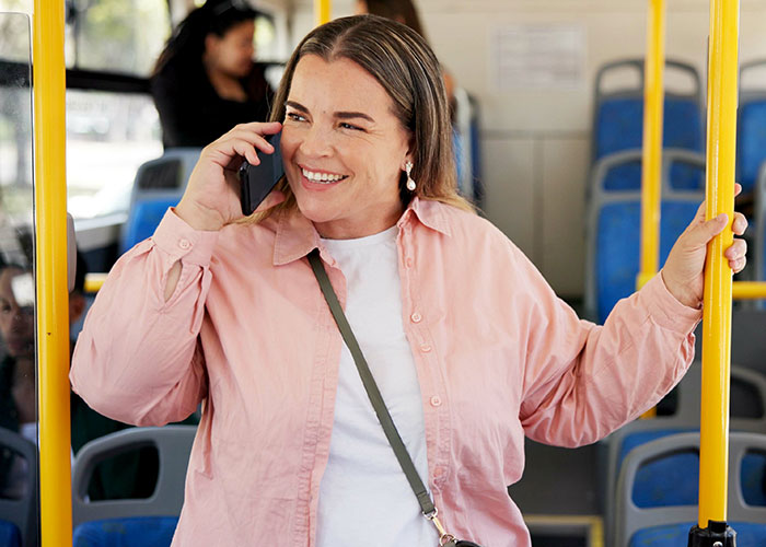 Woman smiling while using phone on public bus, illustrating speakerphone user stories and reactions from others. Woman smiling while using phone on public bus, illustrating speakerphone user stories and reactions from others.