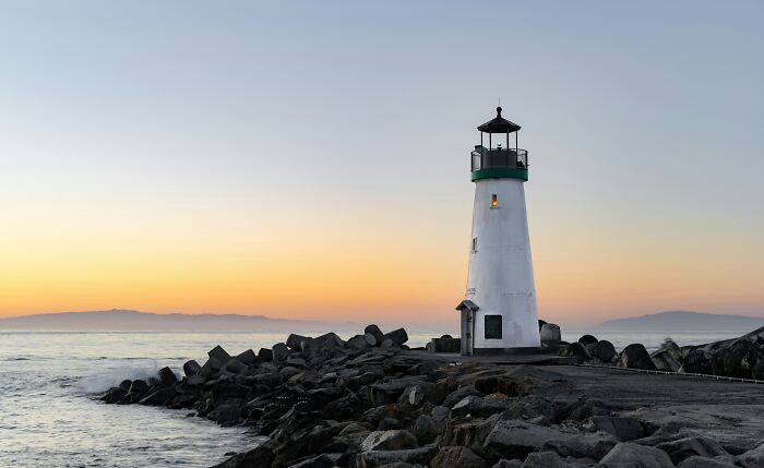 Lighthouse at ocean shore during sunset with rocks in foreground and calm sea, capturing ocean fascination and mystery.