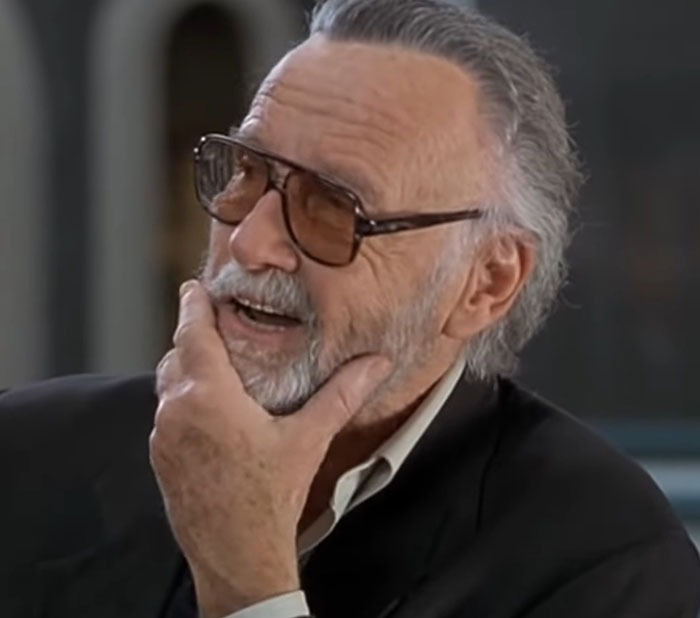 Older man with gray hair and glasses, in a dark suit, smiling during a conversation about encounters with celebrities.