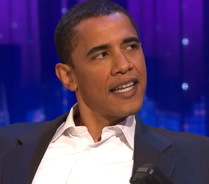 Man in a dark suit and white shirt sharing celebrity encounters during a televised interview with a purple backdrop.