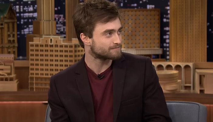 Young man in a dark blazer and maroon shirt sharing a casual moment during a celebrity encounter on a talk show set.