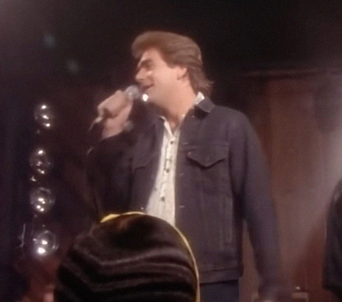Man singing on stage with microphone during celebrity encounter event in a dimly lit venue.