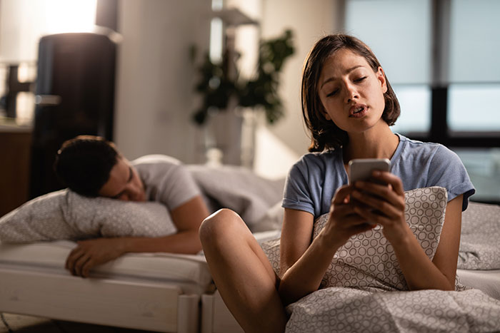 Young woman frustrated scrolling on phone in bedroom while partner lies upset in bed, illustrating life-ruining mistakes.