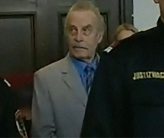 Elderly man in a light suit escorted by officials, representing dark and disturbing Wikipedia pages online topics.