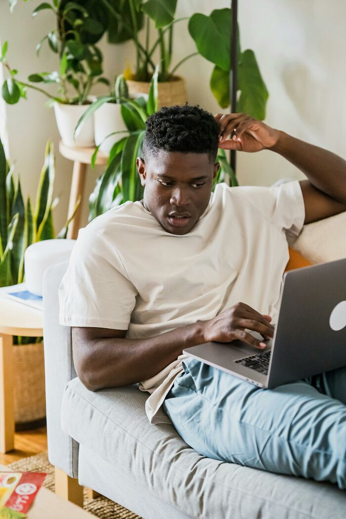 Young man sitting on couch using laptop, looking concerned while exploring disturbing Wikipedia pages and dark rabbit holes online.