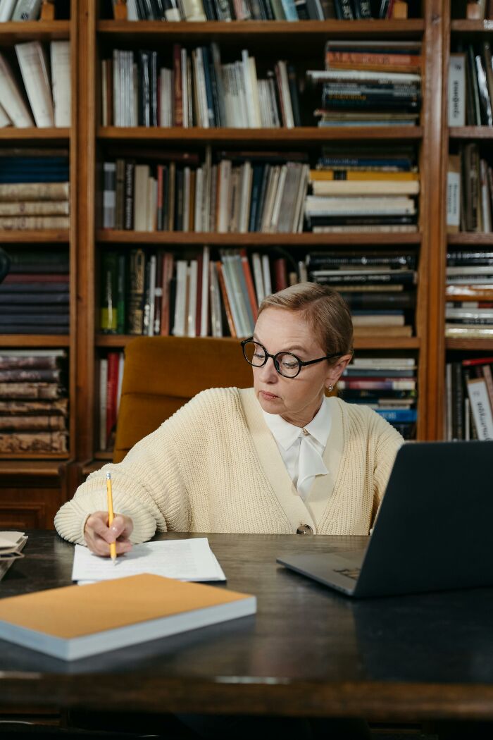 Older woman healthcare worker writing notes at desk with laptop and bookshelf in background, focused on work.
