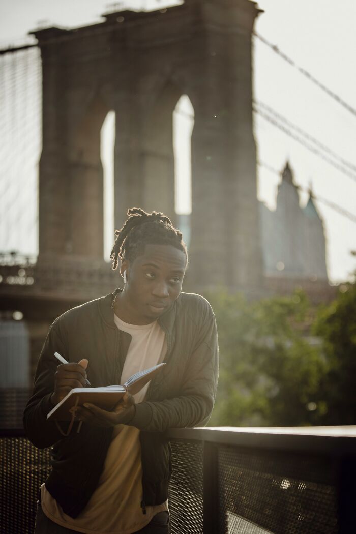 Young person with dreadlocks wearing earbuds and jacket, writing notes near a bridge in urban outdoor setting, unvaccinated healthcare worker.