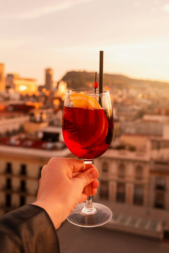 Hand holding a glass of red cocktail with orange slice against a cityscape at sunset, highlighting uncommon popular foods.