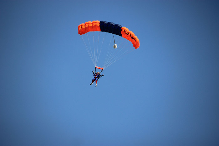 Person skydiving with an orange and black parachute against a clear blue sky in a nightmare survival situation.