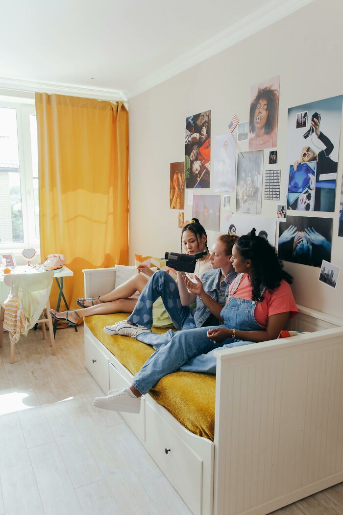 Three friends sitting on a bed in a decorated room, capturing moments with a vintage video camera, childhood rules vibe.