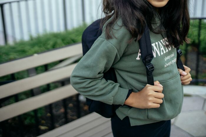 Child holding backpack straps wearing green sweatshirt outdoors near bench focusing on childhood rules concept