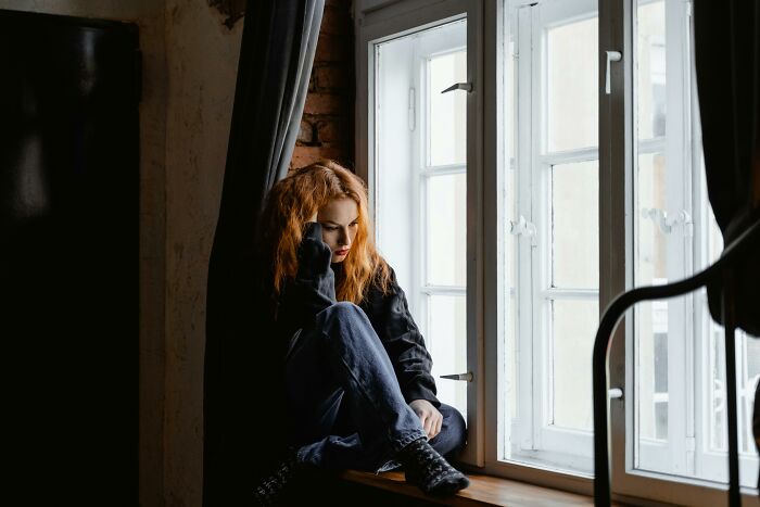 Young woman sitting by a window, deep in thought, reflecting on childhood rules that impact mental health and therapy.