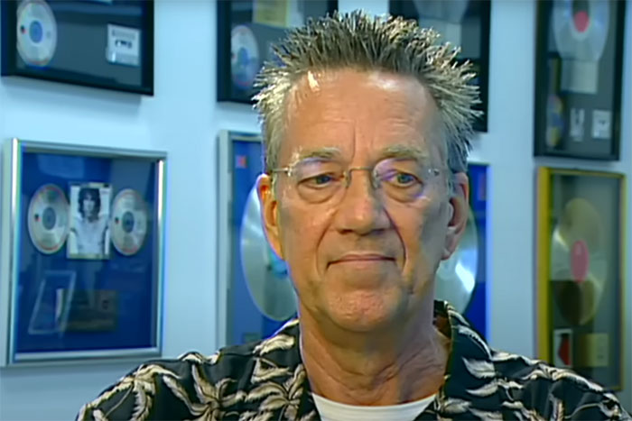 Older man with glasses and spiked hair sitting in a room filled with framed celebrity music awards.