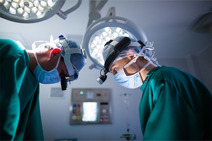 Two surgeons in green scrubs and masks focused during surgery under bright operating room lights.