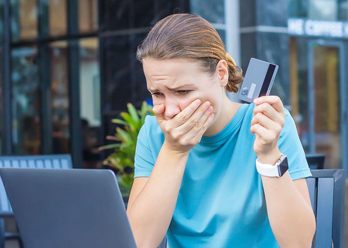 Young woman shocked holding credit card while looking at laptop, illustrating how to ruin your entire life with online stories.