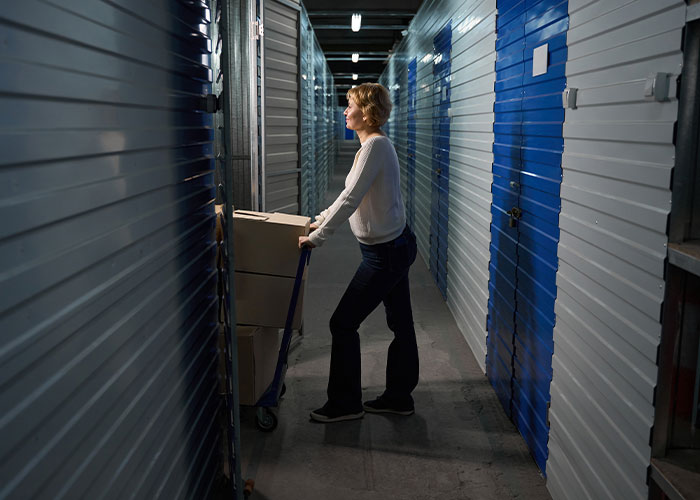 Woman pushing boxes on a dolly in a dimly lit storage unit hallway, illustrating how to ruin your entire life concept.