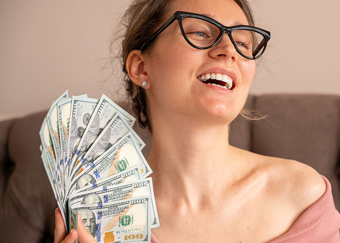 Woman wearing glasses, smiling and holding a fan of hundred-dollar bills representing how to ruin your entire life.