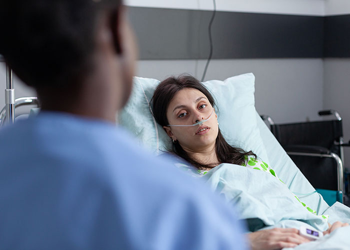 Patient in hospital bed with oxygen tube, appearing weak while a medical professional talks to her, illustrating life challenges.