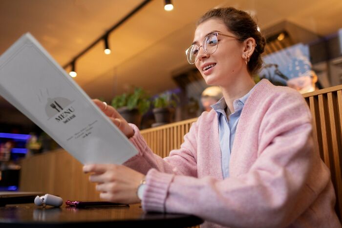 Young woman in glasses reading menu at restaurant, capturing hilariously painful moments of misreading social cues.