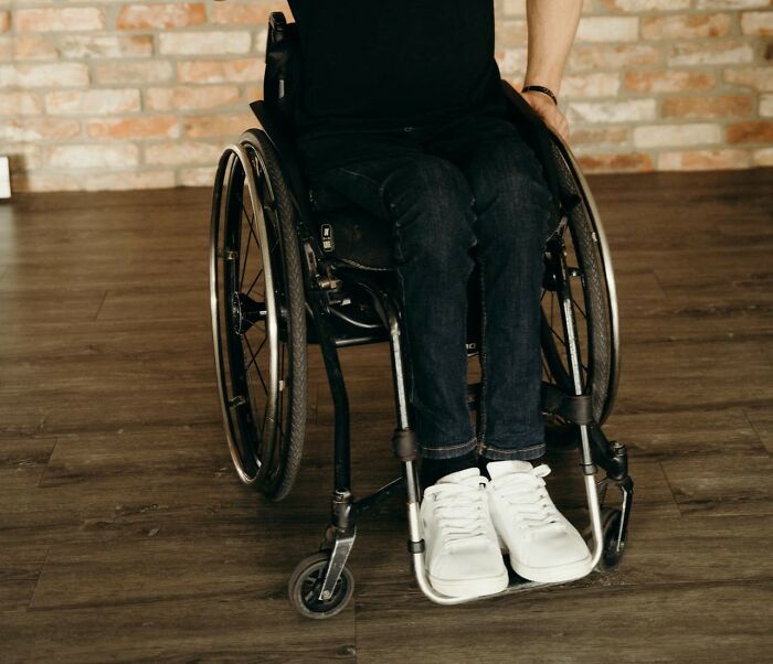 Person sitting in wheelchair on wooden floor, wearing white sneakers and dark jeans, facing brick wall background.