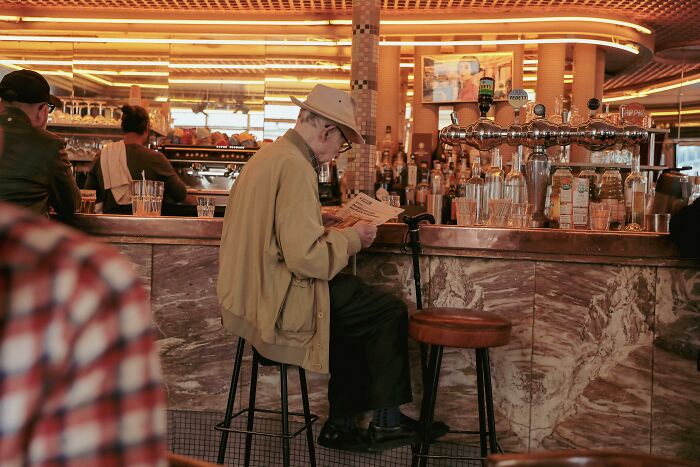 Elderly man sitting alone at a bar counter reading a menu, illustrating a painful table for two moment ending as table for one.