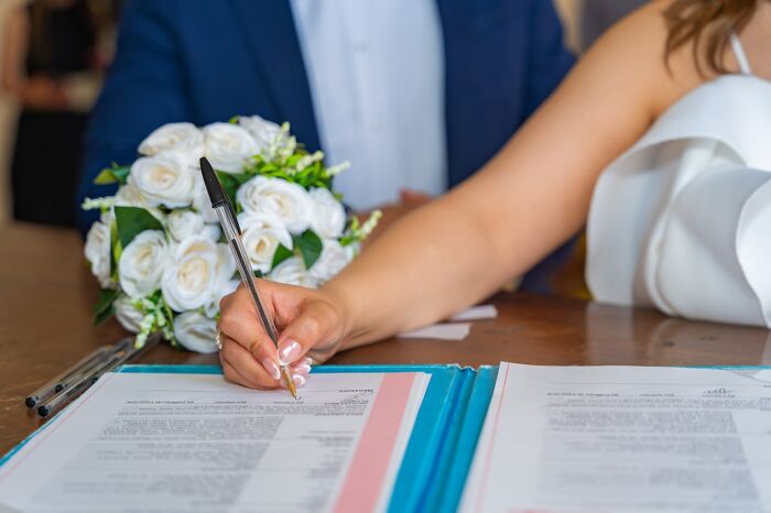 Close-up of a woman signing documents with a bouquet of white flowers nearby, highlighting subtle sexist norms for men.