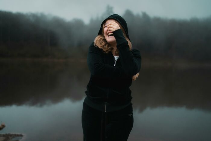 Woman laughing joyfully near a lake, expressing the thrill of karma striking with zero mercy in a misty outdoor setting.