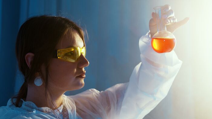 Woman wearing safety glasses carefully examining a flask containing orange liquid, highlighting potentially dangerous situations.