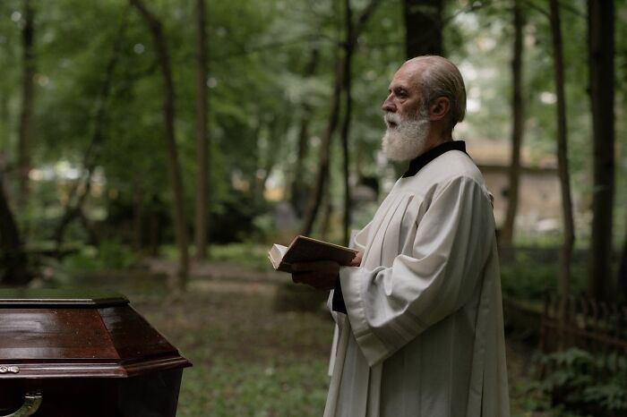 Older priest in white robes holding a book and standing near a coffin during an outdoor funeral ceremony in a forest setting