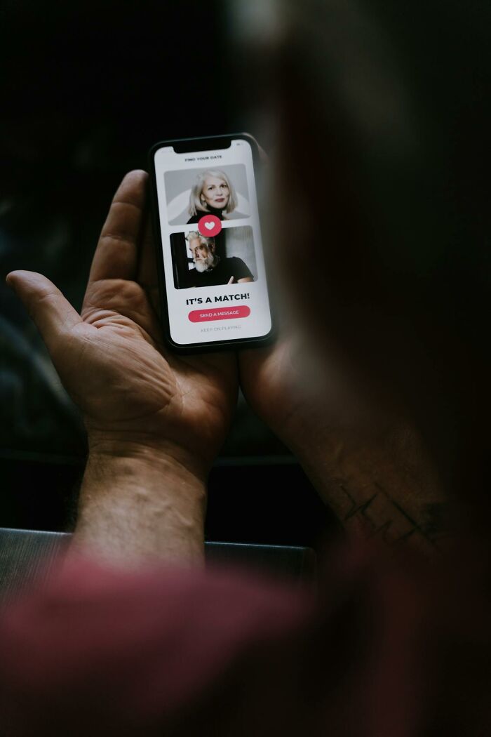 Person holding smartphone with online dating app showing a match notification and heart icon in a dimly lit setting.