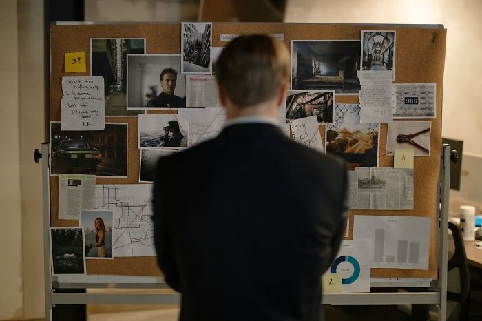 Man in a suit analyzing a true crime investigation board filled with photos, notes, and maps in a dimly lit room.