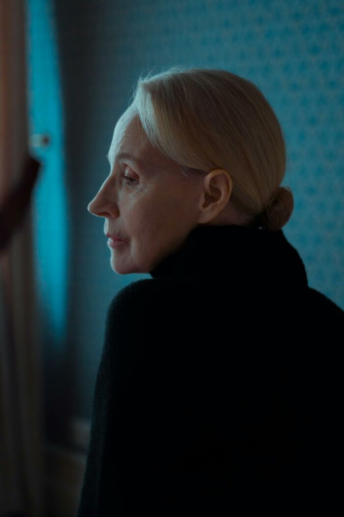 Elderly woman in profile wearing black, reflecting in a dimly lit room, evoking funeral stories and somber mood.