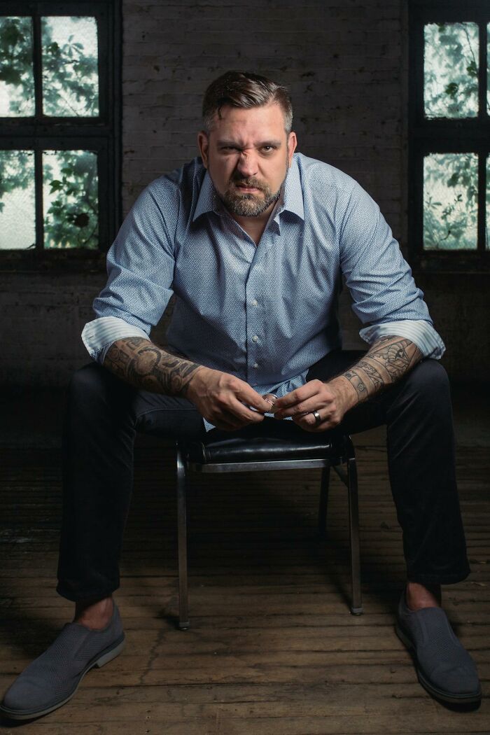 Man with tattoos sitting on a chair in a dim room, looking intensely, representing worst and craziest funeral stories.