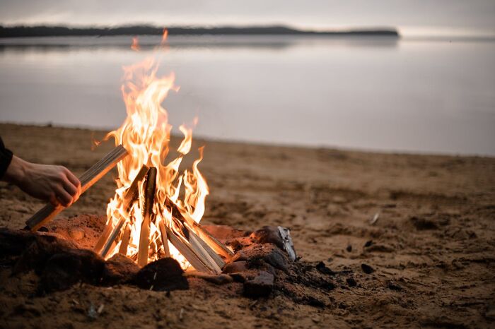 Campfire flames burning brightly on a sandy beach near calm water, symbolizing karma striking with zero mercy.