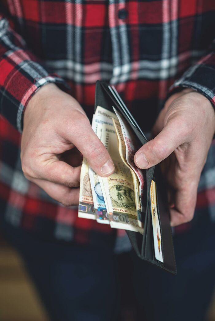 Person in a red plaid shirt holding a wallet with various currency bills visible, illustrating karma concept.