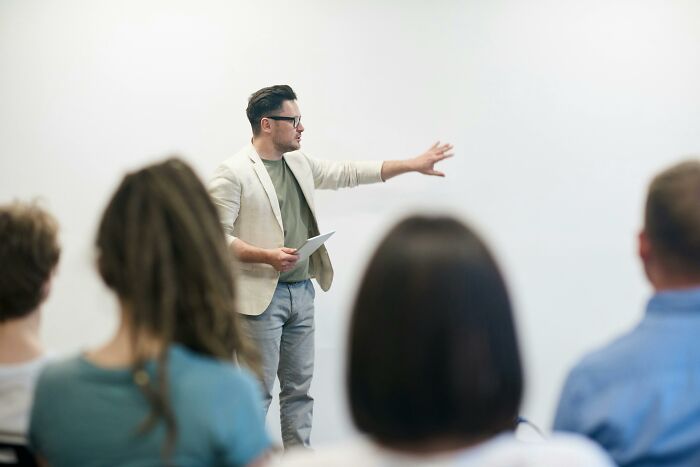 Man in a beige jacket giving a presentation to an audience, illustrating unvaccinated healthcare worker concerns.