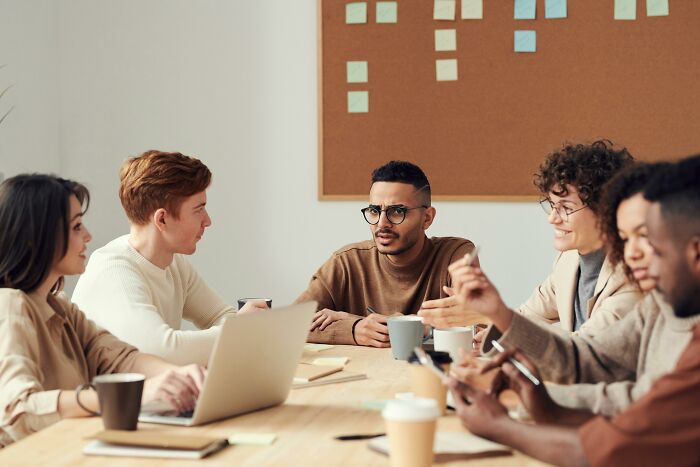 A diverse group of coworkers discussing workplace rules during a meeting, showing visible mixed reactions and engagement.