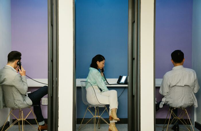 Three office workers sitting in separate phone booths, illustrating over-the-top workplace rules causing annoyance.