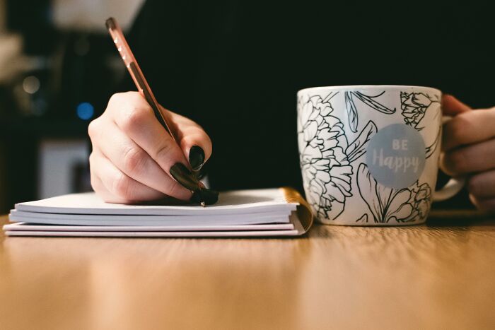 Person with black nail polish writing notes with a pen next to a floral coffee mug, symbolizing an unvaccinated healthcare worker.
